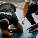 An athlete being assisted by a coach during an indoor training session on a blue floor.